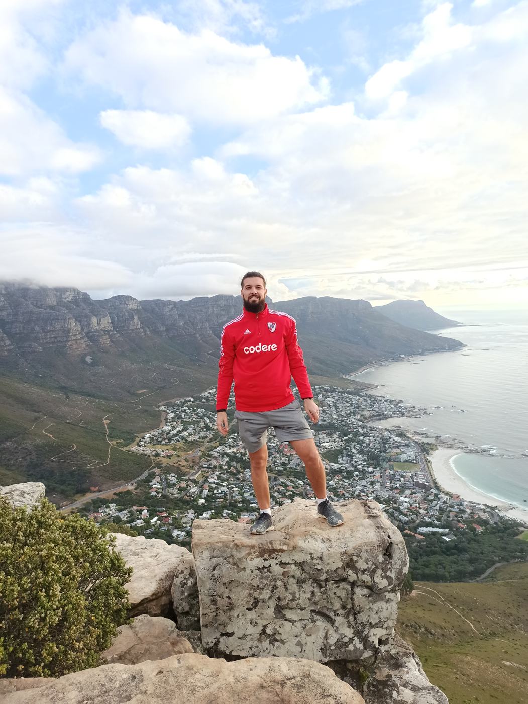 Ismael in a red football jacket standing on a rock above Camps Bay with the mountains and ocean behind