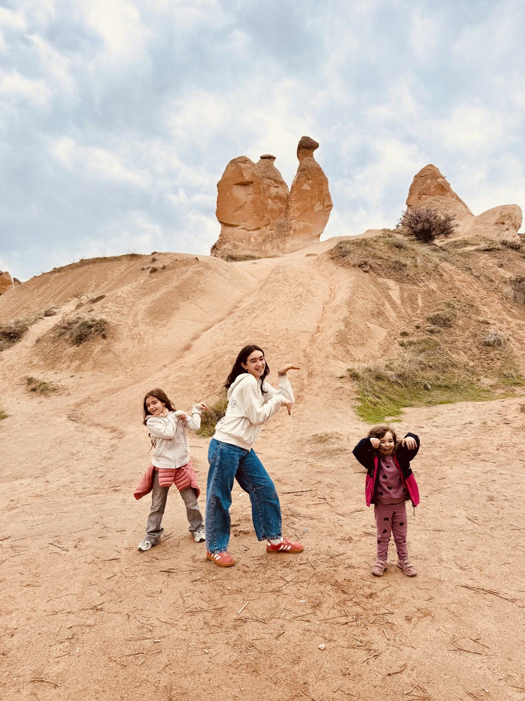 Three girls striking camel poses in front of a camel-shaped rock formation under a cloudy sky