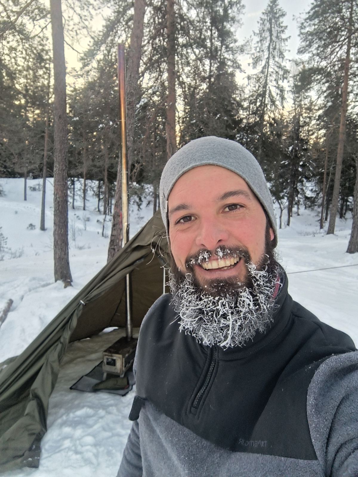 Ismael smiling with a frozen, frosty beard in front of a hot tent in a snowy pine forest