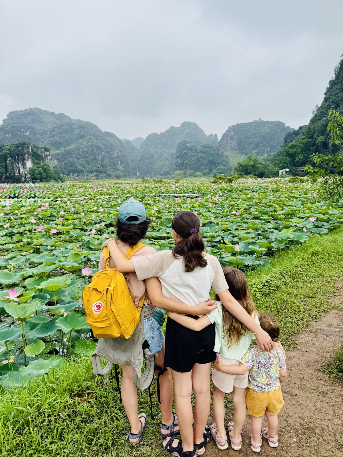 Family arm in arm facing a lotus field and green limestone hills, one with a yellow Kånken backpack