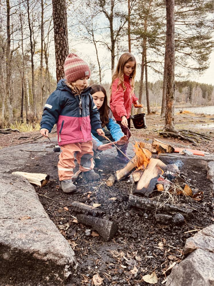 Three girls warming hands and roasting sticks over a campfire by a lake in autumn