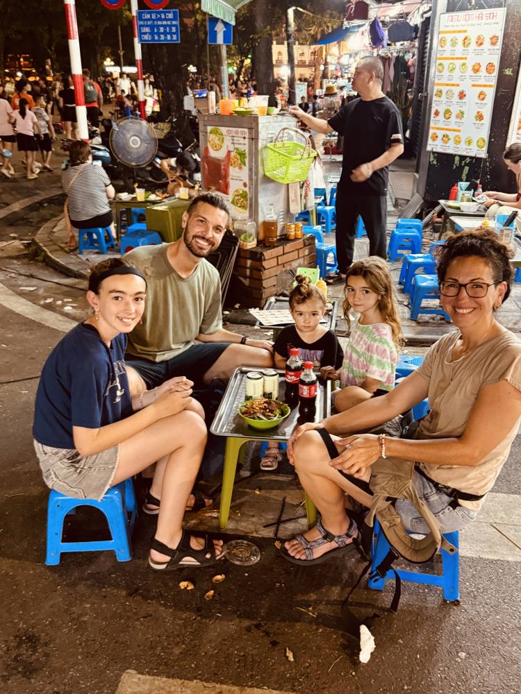 Family at a low table on blue plastic stools at a night street-food corner in Hanoi