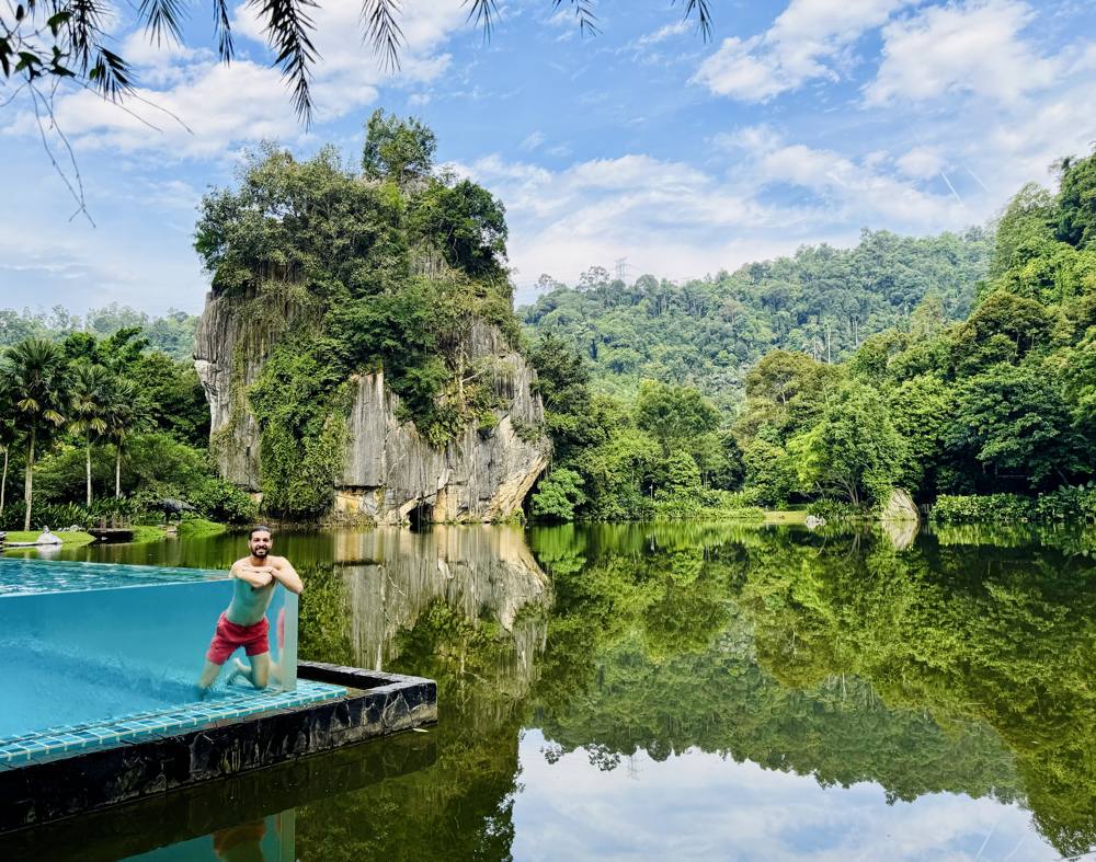 Ismael leaning on the glass edge of a pool facing a lake and a jungle-covered limestone outcrop