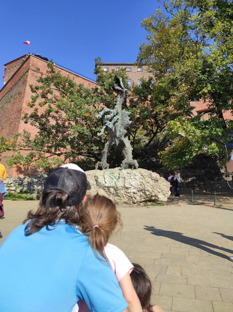 Ismael and his daughter from behind, looking up at the bronze Wawel Dragon below a red-brick castle wall