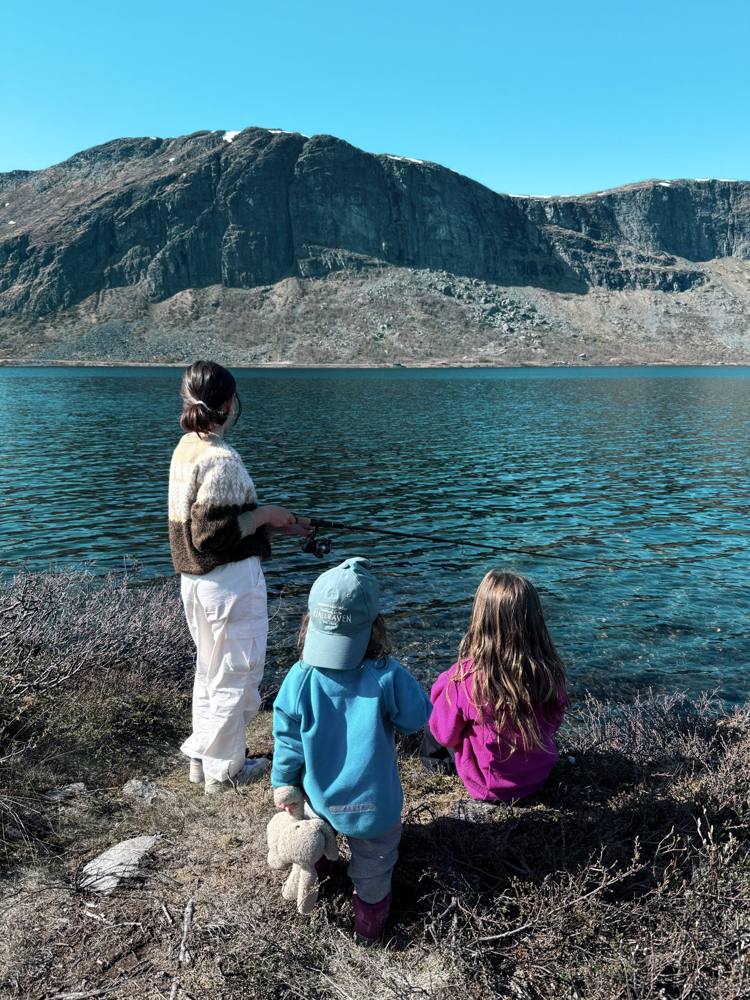 Three children by a turquoise mountain lake, one fishing, under a rocky peak