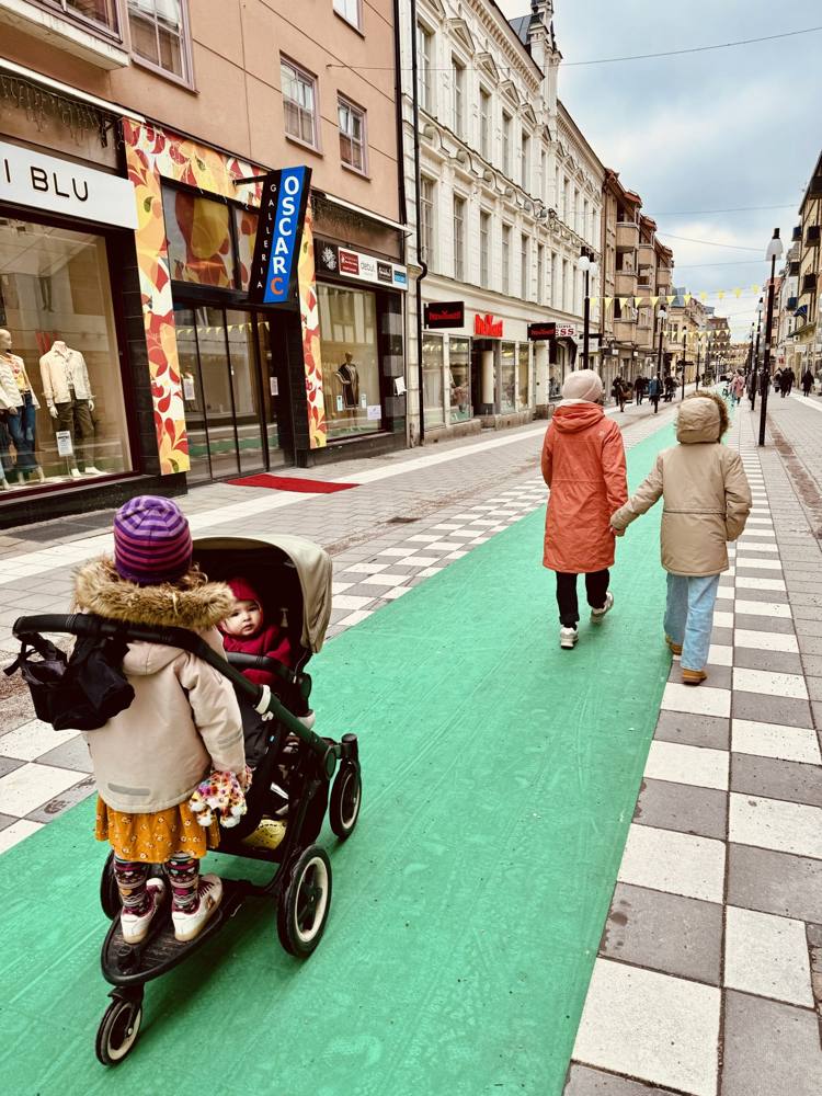 Two girls walking on a green bike lane on a Swedish shopping street, one with a pram
