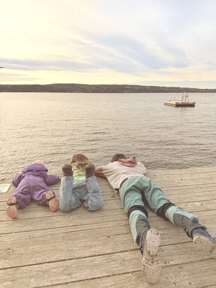 Three people lying on their stomachs on a wooden jetty looking over the Oslofjord at dusk
