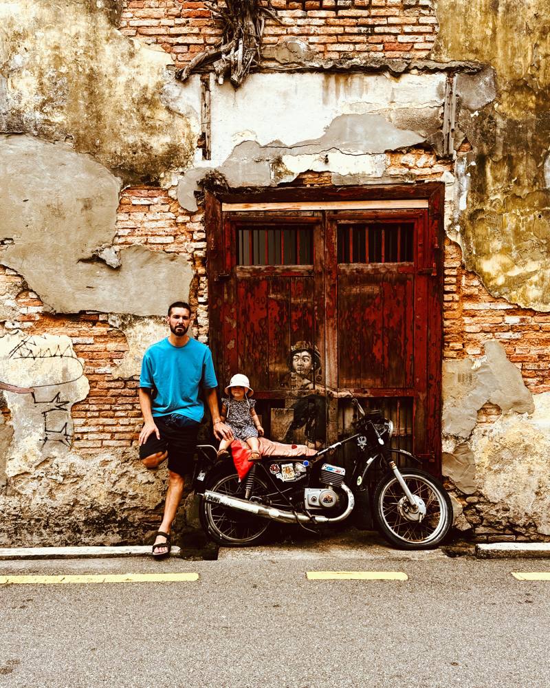 Ismael and a toddler in a sun hat beside a vintage motorbike in front of a weathered brick wall and red door