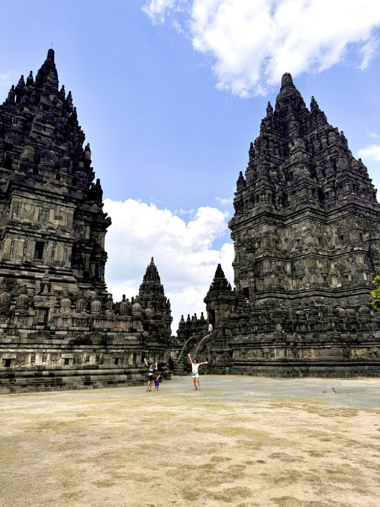 Two tiny figures with arms raised in front of the huge dark spires of Prambanan temple