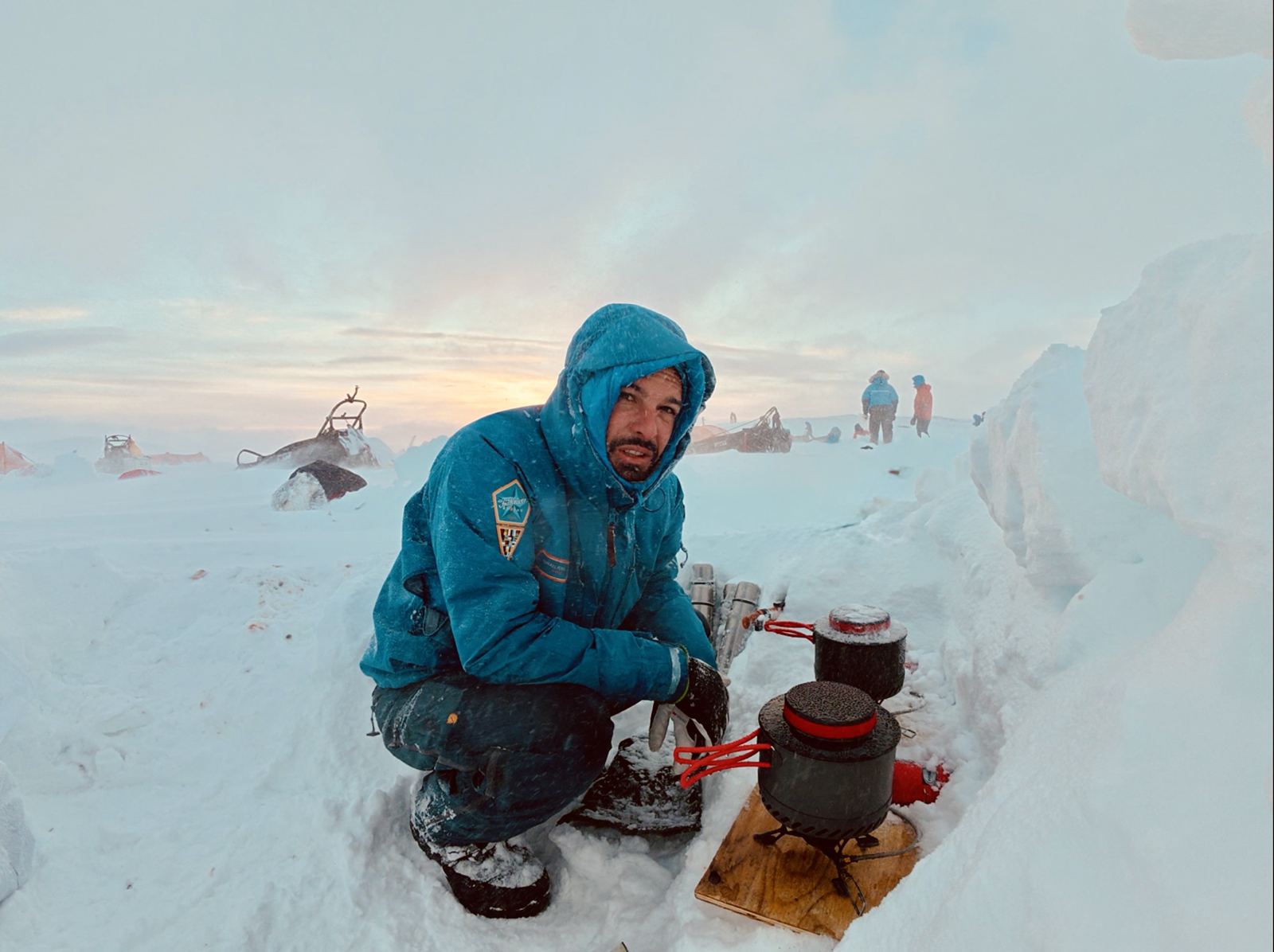Ismael crouching in a snow trench, cooking on a stove in a blowing Arctic storm at sunset