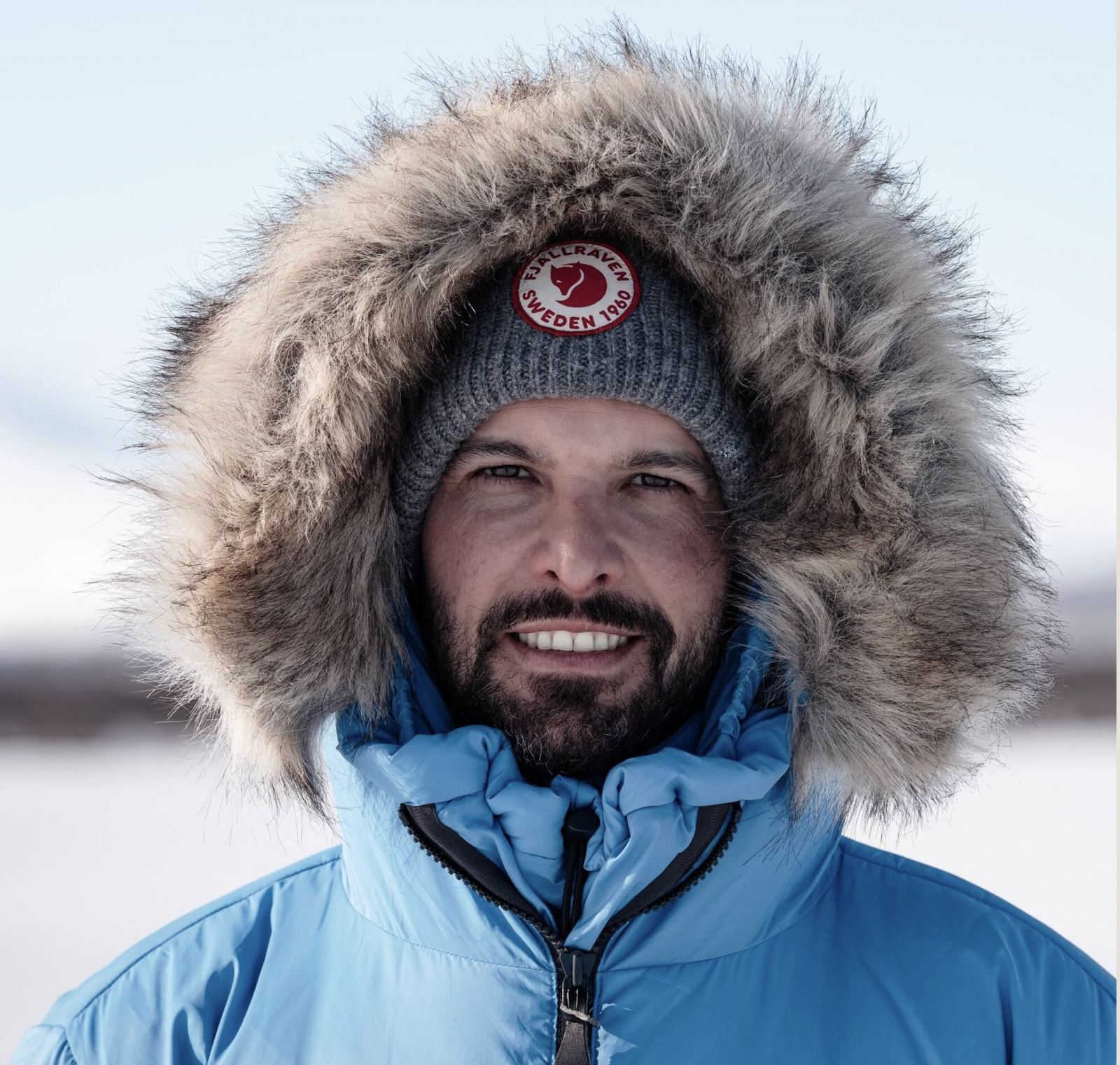 Ismael in a fur-trimmed parka and Fjällräven beanie, smiling in the snow