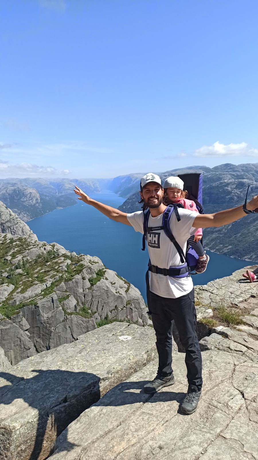 Ismael with arms wide on a cliff above a blue fjord, toddler daughter in a carrier on his back