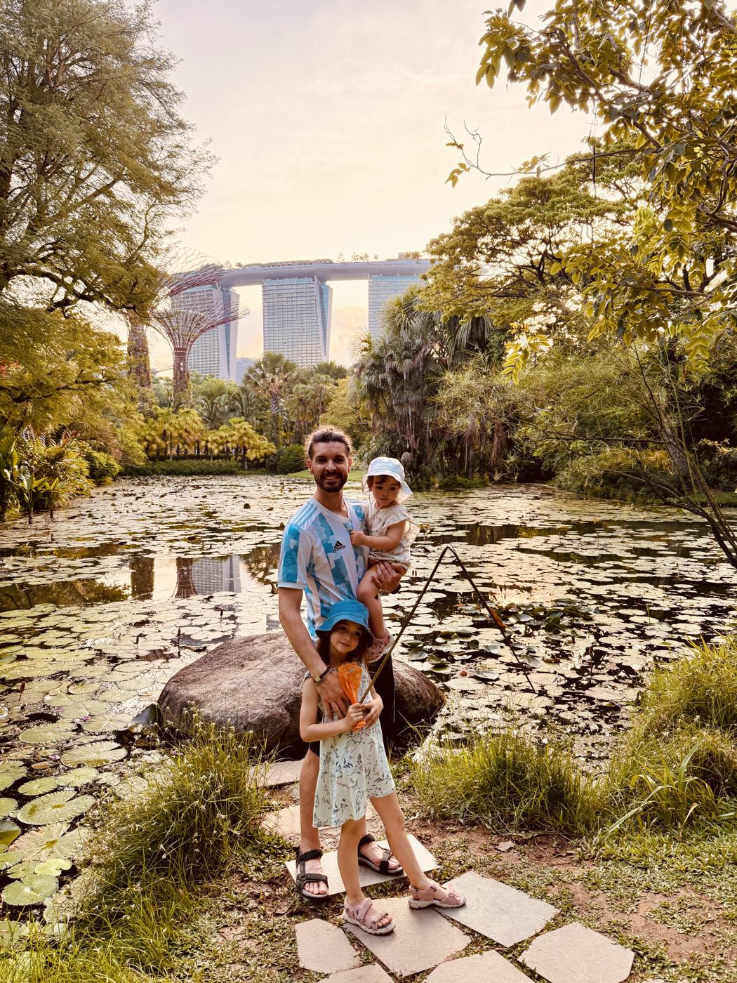 Ismael in an Argentina shirt holding a toddler, with a girl holding a fishing net, beside a lily pond with Marina Bay Sands behind
