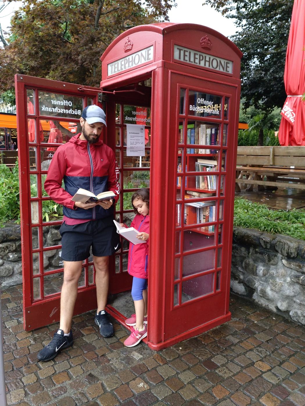 Ismael in a red jacket and his daughter reading books inside a red British-style phone booth turned into a free book exchange