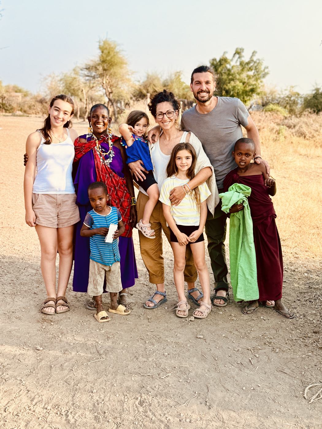 Ismael, his wife and three daughters standing arm in arm with a woman in bright red and purple traditional dress and two village children on a dry dirt road at golden hour