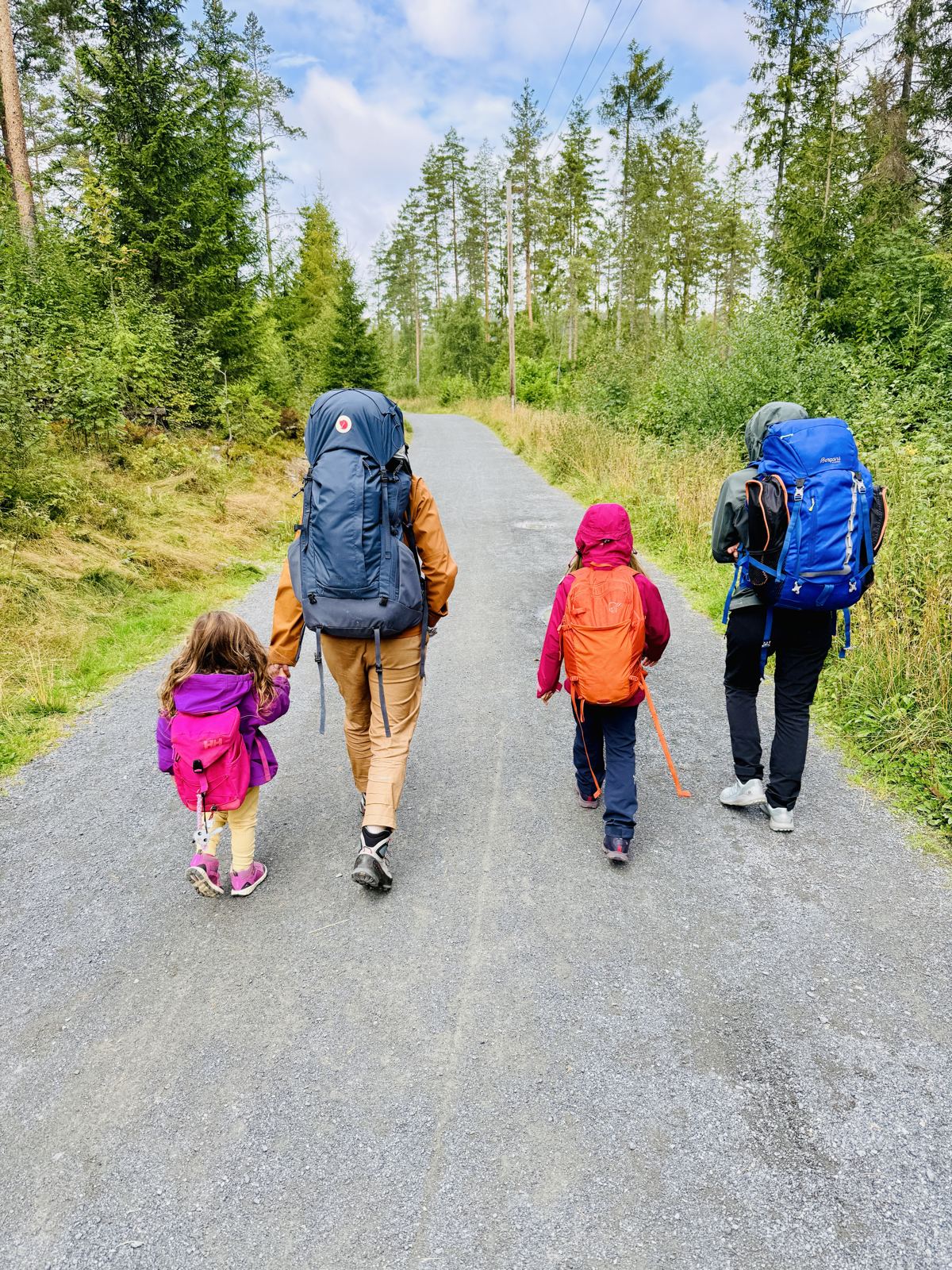 Two adults and two small girls in rain jackets and backpacks walking away on a forest gravel road